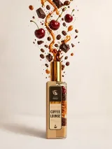 Натюрморт при теплому світлі: флакон Coffee Lounge, латте в керамічній чашці, стиглі вишні у скляній вазочці і обсмажені кавові зерна на бежевому камені